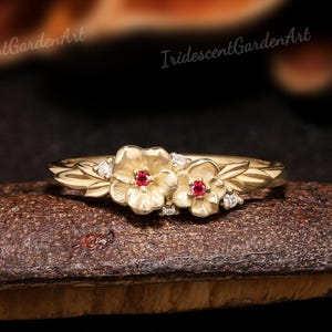 May include: A gold ring featuring two sculpted flower motifs with a central red gemstone in each bloom. The band is adorned with leaf-like details and small clear accent stones. The text IridescentGardenArt is visible in the background.