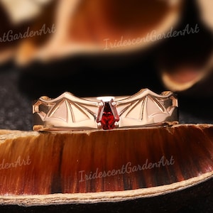 May include: A rose gold ring with a bat wing design and a central, deep red gemstone. The ring is set on a textured, brown surface, with the words "IridescentGardenArt" visible in the background.