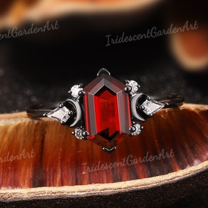 May include: A black ring with a large, rectangular, deep red gemstone as the centerpiece. The ring features smaller white gemstones and crescent moon designs on either side of the main stone. The ring is set against a dark background.