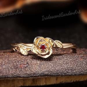 May include: A gold ring featuring a delicate rose design with a small red gemstone at its centre.