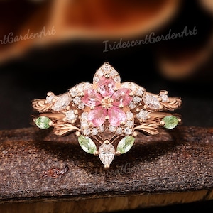 May include: A rose gold ring featuring a central flower motif composed of five pear-cut pink stones, surrounded by small clear accent stones, marquise-cut green stones, and leaf-like metal detailing. The text IridescentGardenArt is visible.