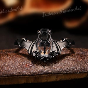 Könnte beinhalten: Ein schwarzer ringförmiger Fledermausring mit detaillierten Flügeln und einem zentralen Stein. Der Ring ist auf einer dunklen, strukturierten Oberfläche platziert, die das gotische Design hervorhebt. Der Ring hat kleine, dunkle Steine.