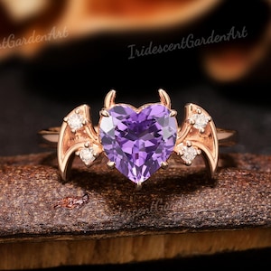 Może przedstawiać: Pierścionek z różowego złota z fioletowym kamieniem szlachetnym w kształcie serca. Pierścionek ma małe rogi i akcenty w kształcie skrzydeł z małymi białymi kamieniami. Widoczny jest tekst "IridescentGardenArt".