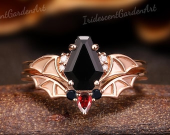 Könnte beinhalten: Ein Ring aus Roségold mit einem schwarzen Onyx-Edelstein in einem Fledermausflügel-Design. Der Ring ist mit kleineren Diamanten und roten Edelsteinen besetzt.