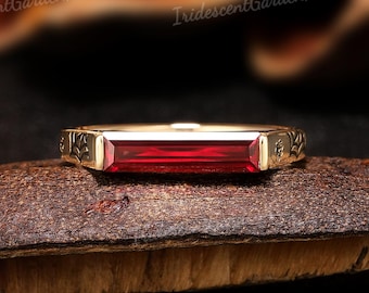 May include: A gold-colored ring with a rectangular, deep red gemstone. The ring has decorative carvings on the sides and is set on a brown, textured surface. The ring's design is elegant and eye-catching.
