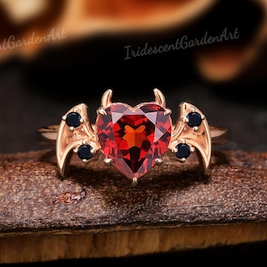 Op de afbeelding: Een roségouden ring met een hartvormige, gefacetteerde rode edelsteen. De ring heeft kleine zwarte edelstenen en vleugelachtige accenten aan weerszijden van het hart. De ring ligt op een donker, getextureerd oppervlak.