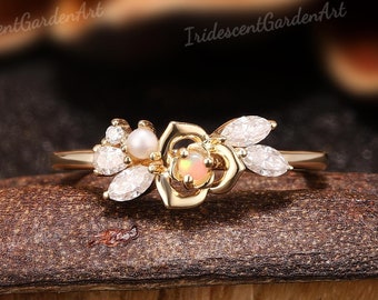 May include: A gold ring with a delicate rose design featuring a small opal gemstone and white pearls. The ring is set with small diamonds and has a delicate floral design.