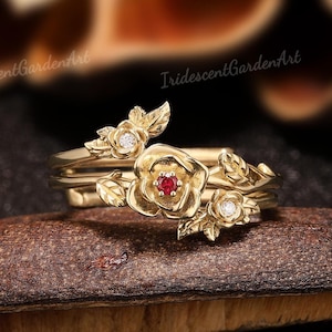 以下が含まれることがあります： 金色のリングセット。中央のリングは、赤い宝石を中央に配したバラのデザインで、小さな花と葉のアクセントで囲まれています。リングセットは茶色の表面に展示されています。