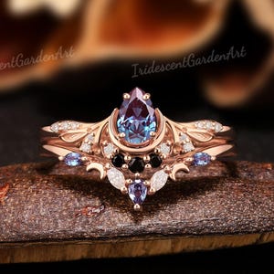 May include: A rose gold ring featuring a central pear-cut color-changing stone, accented with smaller round black stones, marquise-cut white stones, and light blue pear-cut stones. The band has a curved, layered design with intricate metalwork.