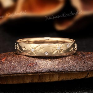 Puede incluir: Un anillo dorado con un diseño de estrellas y puntos, engastado con pequeñas piedras preciosas transparentes. El anillo se muestra sobre una superficie marrón texturizada con un fondo borroso. El texto "IridescentGardenArt" es visible.