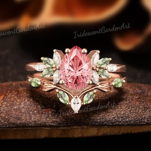 May include: A rose gold ring set featuring a pear-shaped pink gemstone as the centerpiece, surrounded by white and green accent stones. The ring has a floral design with delicate leaf details. The brand name "IridescentGardenArt" is visible.