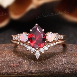 May include: A pear-shaped red gemstone engagement ring with a delicate band of smaller diamonds and two opal stones. The ring is set in rose gold.