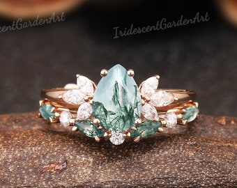 May include: A rose gold engagement ring set with a pear-shaped green moss agate gemstone surrounded by a halo of smaller clear gemstones. The band is adorned with smaller green gemstones.