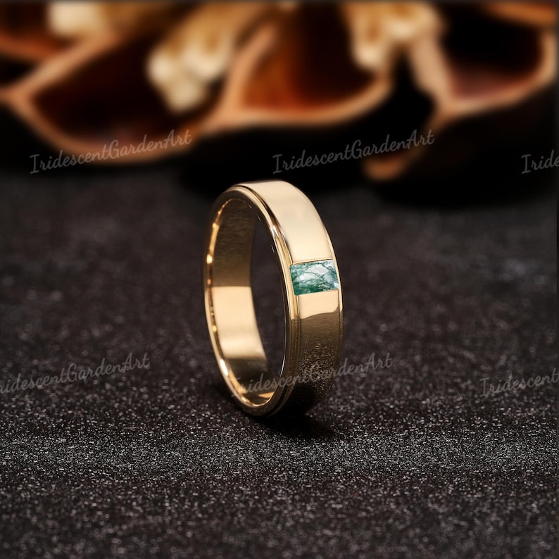 May include: A gold-colored ring with a rectangular, green stone inlay. The ring has a flat band design and a polished finish. The stone appears to be moss agate. The ring is displayed on a dark, textured surface.