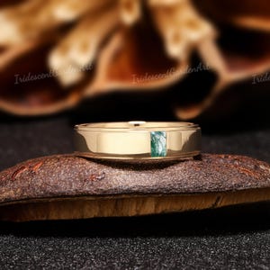 May include: A gold-colored ring with a rectangular green stone inlay. The ring is set on a dark, textured surface, with a blurred background. The ring's band is smooth and polished.
