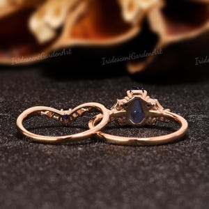 May include: Two rose gold rings. One ring features a large, dark blue, teardrop-shaped gemstone. The second ring is a curved band with small blue gemstones. The rings are set against a dark background.