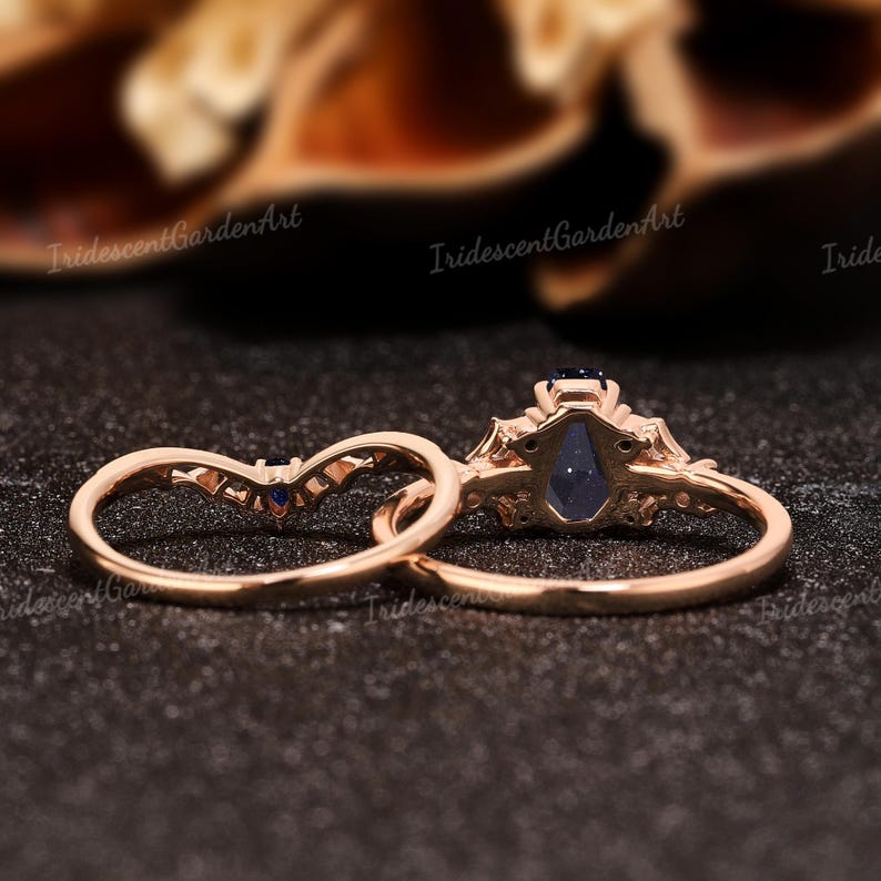 May include: Two rose gold rings. One ring features a large, dark blue, teardrop-shaped gemstone. The second ring is a curved band with small blue gemstones. The rings are set against a dark background.