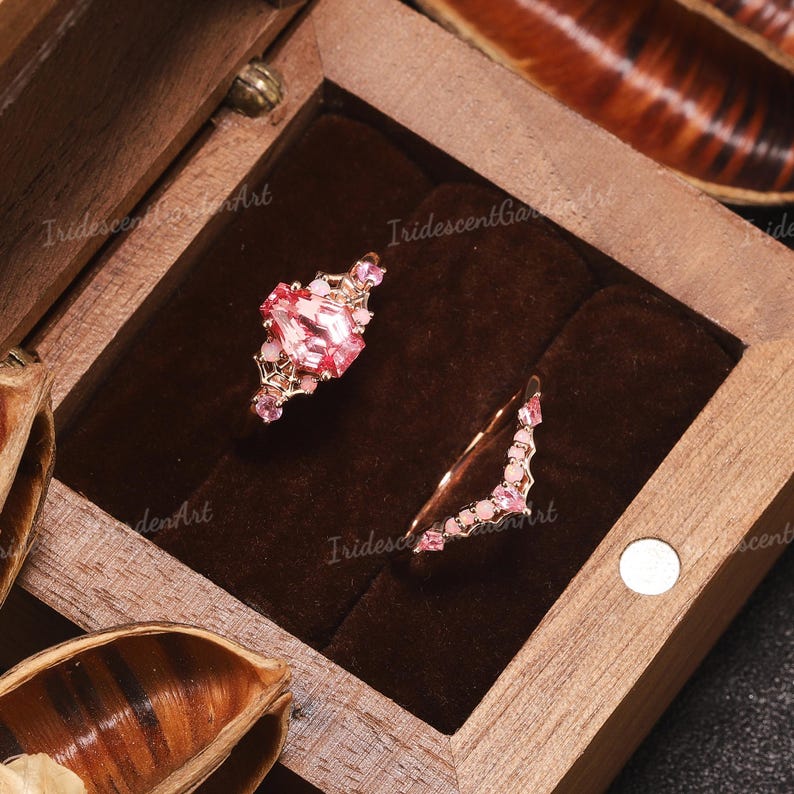 May include: A two-piece ring set featuring a rose gold band with a central coffin-shaped pink gemstone and smaller pink accent stones, paired with a curved band adorned with a row of small pink stones and round pink cabochons.