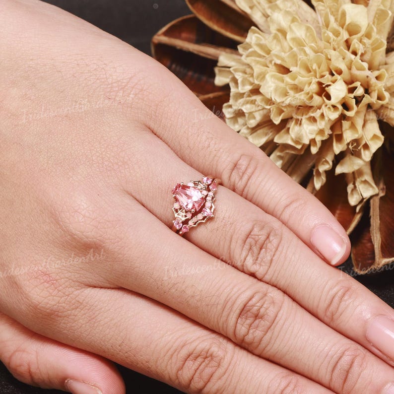 May include: A rose gold ring featuring a central pear-shaped pink gemstone surrounded by smaller pink stones in a delicate, intricate setting. The band is thin and metallic, showcasing a refined design with multiple small, sparkling pink accents.