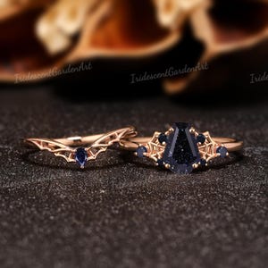 May include: Two rose gold rings. One ring has a curved band with a blue gemstone. The other ring has a large, dark blue, rectangular gemstone with smaller blue gemstones and decorative accents. The rings are on a dark surface.