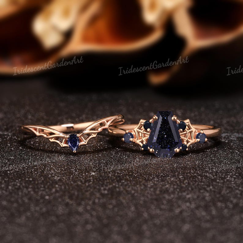 May include: Two rose gold rings. One ring has a curved band with a blue gemstone. The other ring has a large, dark blue, rectangular gemstone with smaller blue gemstones and decorative accents. The rings are on a dark surface.
