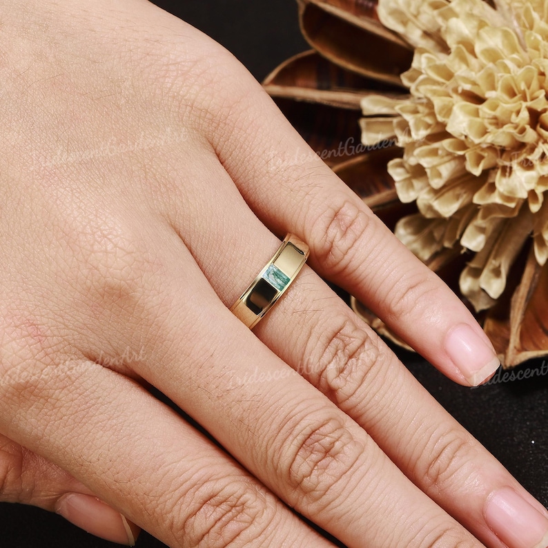 May include: A gold-colored ring with a rectangular, light-blue gemstone set in the center. The ring is on a finger, with a blurred background of a flower and dark surface.