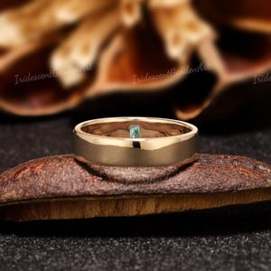 May include: A gold-colored ring with a small, square, light-green gemstone set in the center. The ring has a smooth, polished finish and a wide band. The ring is displayed on a brown, textured surface. The text "IridescentGardenArt" is visible.