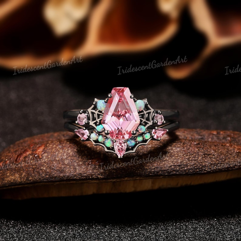 May include: A black metal ring set featuring a central coffin-cut pink gemstone, accented with smaller pink stones and round iridescent opals. The band design incorporates a spider web motif surrounding the stones, creating a detailed, layered appearance.
