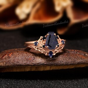 May include: Rose gold ring with a dark blue coffin-shaped stone as the centerpiece, surrounded by smaller dark blue stones. The ring has an intricate filigree design and is set on a dark brown surface.