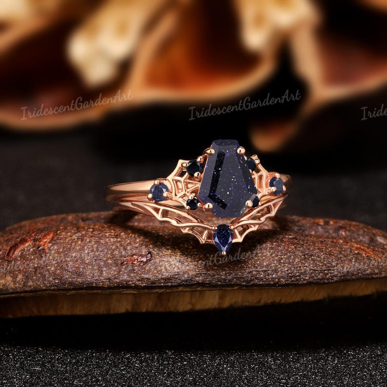 May include: Rose gold ring with a dark blue coffin-shaped stone as the centerpiece, surrounded by smaller dark blue stones. The ring has an intricate filigree design and is set on a dark brown surface.