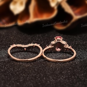May include: A rose gold ring set consisting of a curved band and a main ring featuring a pear-shaped pink gemstone at the center, accented with smaller stones along the band. The metal has a warm, metallic finish and the design is delicate.
