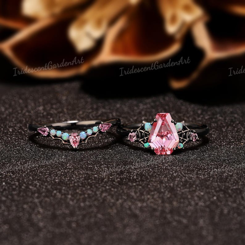 May include: A black metal ring set featuring a coffin-cut pink center stone flanked by spider web detailing and small iridescent opal and pink accents, paired with a matching chevron-shaped band adorned with opals and pink stones.