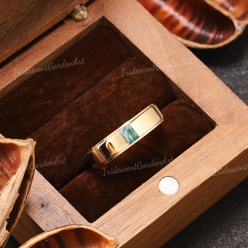 May include: A gold ring with a rectangular, light green stone inlay. The ring is displayed inside a wooden box with a dark brown velvet lining. The ring has a polished, reflective surface.