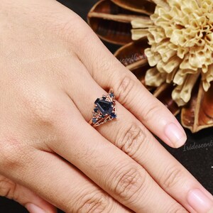 May include: A rose gold ring featuring a dark blue, square-cut gemstone at the center, surrounded by smaller blue stones. The ring has an intricate, openwork design. The ring is on a person's finger.
