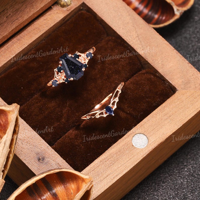 May include: Two rose gold rings displayed in a wooden box. One ring features a large, dark blue gemstone with smaller blue accents. The other ring has a wing-like design with a small blue stone.