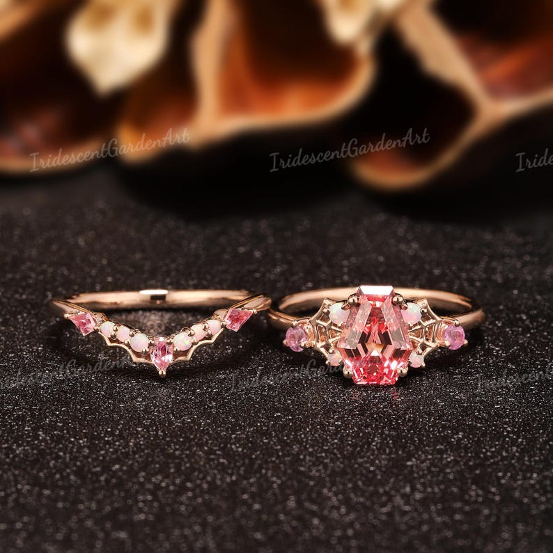 May include: A rose gold ring set featuring a central coffin-cut pink gemstone flanked by small round opals and pink stones, paired with a matching chevron-shaped band adorned with pink stones and opals. The metal has a delicate filigree design.