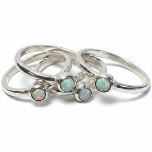 May include: A collection of three silver rings, each featuring a single round, bezel-set opal gemstone. The opals display iridescent flashes of blue, green, and orange light. The bands are smooth, polished silver, arranged in a stacked formation.