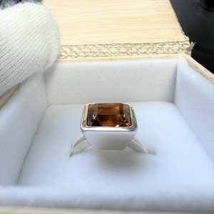 May include: A silver ring with a large, rectangular, brown gemstone set in a square bezel. The ring is in a white box.
