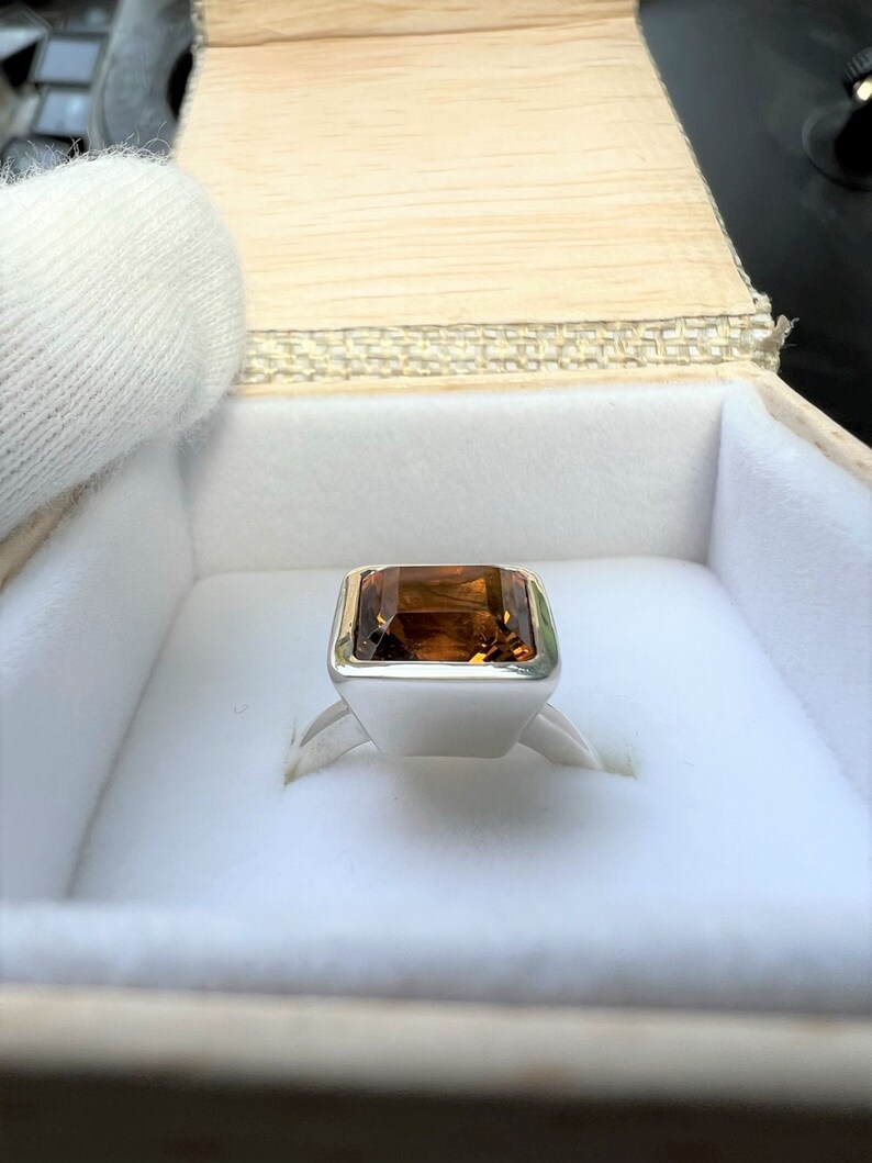 May include: A silver ring with a large, rectangular, brown gemstone set in a square bezel. The ring is in a white box.