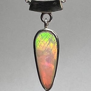 Puede incluir: Un collar con colgante de plata con una piedra preciosa en forma de lágrima que muestra colores vibrantes: verde, amarillo, naranja y rosa. La piedra está engastada en plata y cuelga de una cadena de plata.