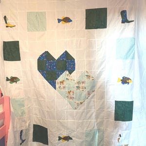 May include: A white patchwork quilt featuring two large central heart shapes, one made of dark green and blue patterned fabric and the other of light blue gingham and farm animal print. The border includes squares of green, gingham, and fish and boot motifs.