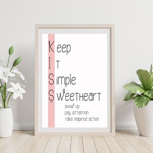 May include: A framed print with a pink stripe on the left side. The print features the acronym KISS with the words Keep It Simple Sweetheart written vertically. Below the acronym, the words show up, pay attention, take inspired action are written in black.
