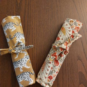 May include: Two fabric utensil rolls. One is mustard yellow with white leopard print, tied with a matching bow. The other is cream-colored with a floral pattern in red, orange, and green, tied with a coordinating bow. Both are on a wooden surface.