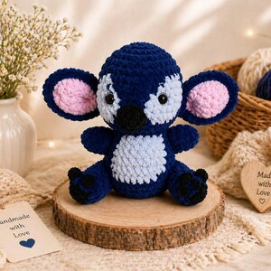 Puede incluir: Un juguete de peluche de ganchillo azul y blanco hecho a mano con el interior de las orejas rosa, sentado sobre una rodaja de madera. El juguete tiene ojos negros y una nariz negra. Una etiqueta que dice "Handmade with Love" est&aacute; al lado.