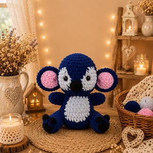 Puede incluir: Un koala de ganchillo azul y blanco hecho a mano con orejas internas rosas y detalles negros. El peluche est&aacute; sentado sobre una estera redonda tejida. El koala tiene una barriga blanca y est&aacute; rodeado de objetos decorativos.