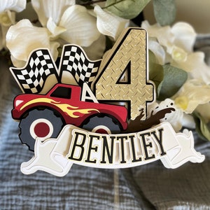 May include: A red monster truck cake topper with a checkered flag, a gold number four, and the name 'Bentley' on a white banner. The truck has flames on the side and is driving through mud.