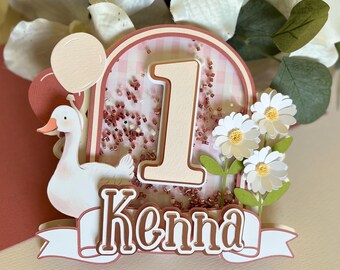 May include: A pink and white birthday cake topper with a white goose, a pink and white gingham background, a large number one, and the name "Kenna" in brown lettering.