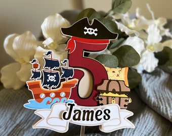 May include: A pirate-themed cake topper featuring a red number 5 with a pirate ship, treasure chest, and a banner that says "James". The topper includes a pirate hat with a skull and crossbones.