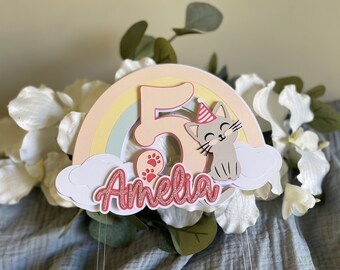 Op de afbeelding: Een pastelkleurige regenboog caketopper met een grijze kat die een rood-witte feestmuts draagt, een roze nummer 5 en de naam Amelia in roze glitterletters.