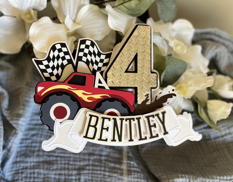 May include: A red monster truck cake topper with a checkered flag, a gold number four, and the name 'Bentley' on a white banner. The truck has flames on the side and is driving through mud.